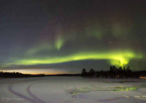 Die untergegangene Sonne schickt etwas Restlicht an den nördlichen Horizont. Ein Polarlichtbogen über dem Fluss Ounasjoki sorgt zusätzlich für eine besondere Lichtstimmung.